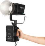 The NANLITE V Mount battery handle with 4-pin XLR connector is in the hand and powers the Forza 150 LED video light, a professional handheld light for use with a 14.8V battery.