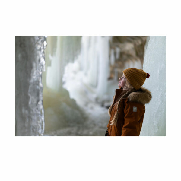 A person in a brown fur coat and yellow knitted jacket stands in an icy cave looking up at the frozen walls - shot with a Sigma 50mm f/1.2 DG DN Art lens on a Sony E.