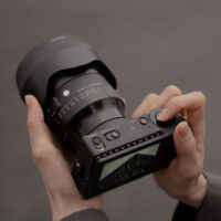 A person is holding a Sigma 50mm f/1.2 DG DN Art lens (Sony E) in both hands - one adjusting the lens, the other using the controls; the camera's display and buttons are visible.