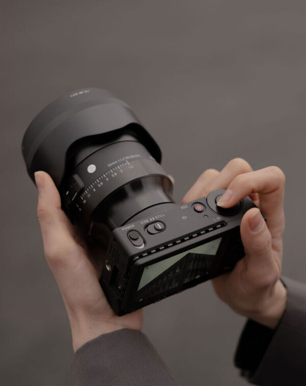 A person is holding a Sigma 50mm f/1.2 DG DN Art lens (Sony E) in both hands - one adjusting the lens, the other using the controls; the camera's display and buttons are visible.