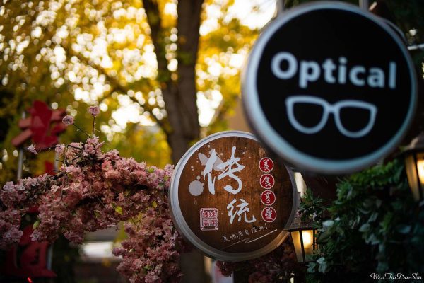 Viltrox MF 85mm f/1.8 PFU RBMH -objektiivilla Sonylle kuvattu kiinalaisilla kirjaimilla koristeltu puinen kyltti on vaaleanpunaisten kukkien kehystämä, kun taas pyöreä optinen kyltti silmälaseineen erottuu auringonvalossa keltaisine lehvästöineen puiden edessä.