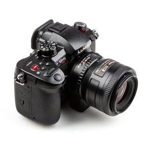 Valkoisella taustalla näkyy musta Panasonic Lumix GH5 -kamera, jossa on erilaisia painikkeita ja valitsimia ja joka on varustettu Viltrox Nikon F-M1 -objektiivin kiinnityssovittimella.