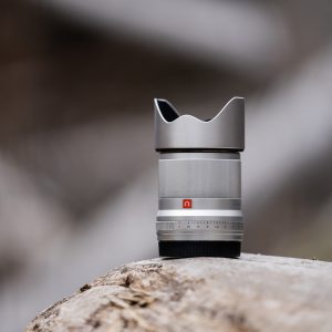 Viltrox AF 33mm f/1.4 -objektiivi, Nikon Z (hopea), jossa on terälehden muotoinen huppu, seisoo pystyasennossa puunrungon päällä pehmeästi sumeaa luonnollista taustaa vasten.