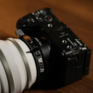 Lähikuva mustasta Sony-kamerasta, johon on kiinnitetty suuri valkoinen objektiivi Viltrox PL-E -sovittimella, puupinnalle asetettuna. Kameran painikkeet, valitsimet ja tuotemerkki ovat selvästi näkyvissä.