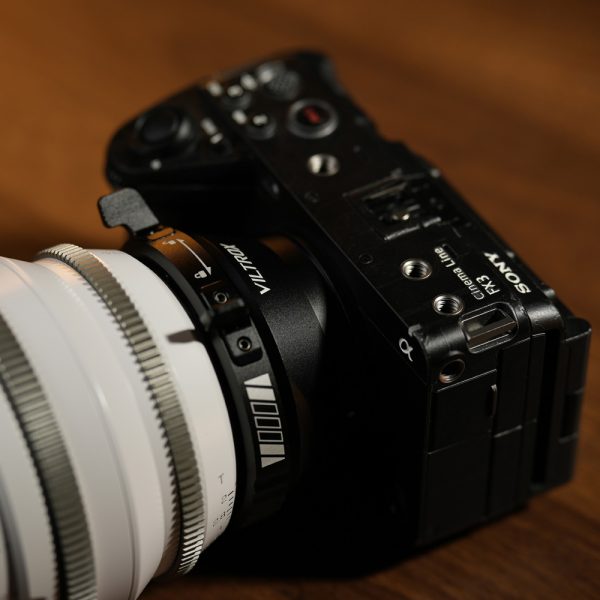 Lähikuva mustasta Sony-kamerasta, johon on kiinnitetty suuri valkoinen objektiivi Viltrox PL-E -sovittimella, puupinnalle asetettuna. Kameran painikkeet, valitsimet ja tuotemerkki ovat selvästi näkyvissä.
