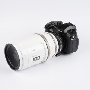 Musta Panasonic Lumix GH6 -kamera, jossa on suuri valkoinen Viltrox 100mm T2.0 -kino-objektiivi, on kiinnitetty Viltrox PL - MFT -sovittimella, ja kaikki on asetettu valkoista taustaa vasten.