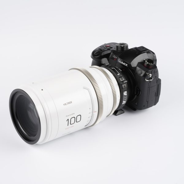 Musta Panasonic Lumix GH6 -kamera, jossa on suuri valkoinen Viltrox 100mm T2.0 -kino-objektiivi, on kiinnitetty Viltrox PL - MFT -sovittimella, ja kaikki on asetettu valkoista taustaa vasten.