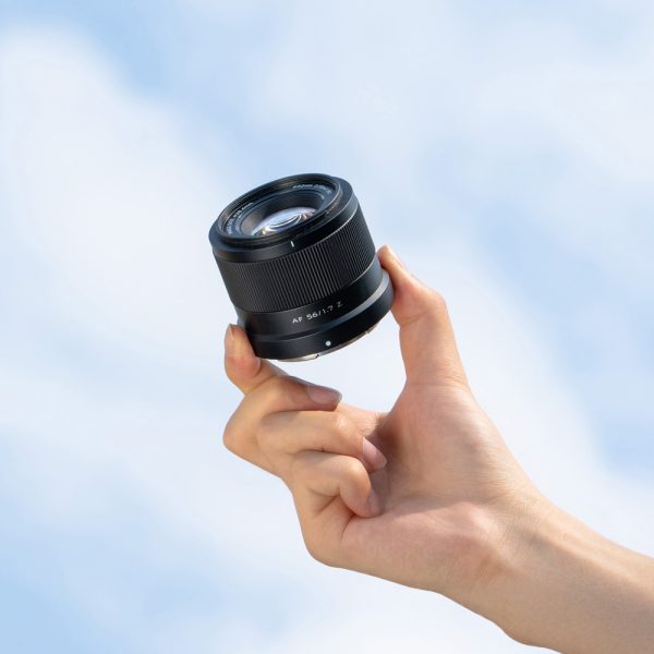 Käsi pitää ylhäällä Viltrox AF 56mm f/1.7 -objektiivi, Nikon Z -objektiivia kirkkaan sinistä taivasta vasten, jonka taustalla on pehmeät valkoiset pilvet.