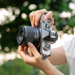 Henkilö pitelee hopean ja mustan väristä Viltrox AF 56mm f/1.7 -objektiivi for Nikon Z -kameraa vaakatasossa ulkona, vaaleat kynnet näkyvät ja taustalla on sumea vihreä tausta.