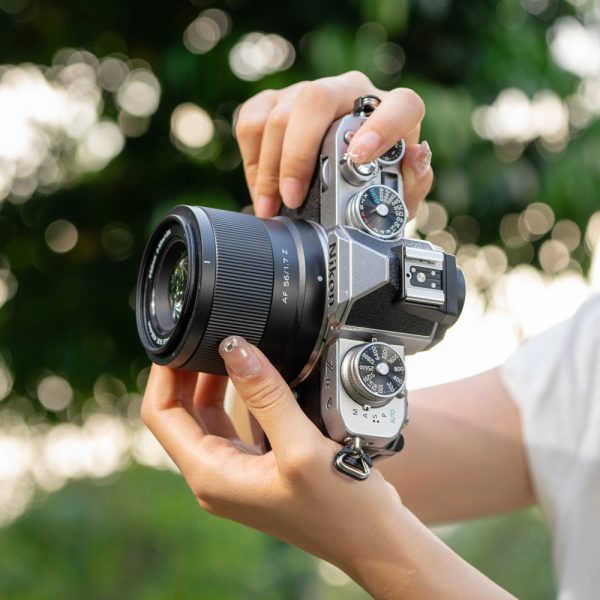 Henkilö pitelee hopean ja mustan väristä Viltrox AF 56mm f/1.7 -objektiivi for Nikon Z -kameraa vaakatasossa ulkona, vaaleat kynnet näkyvät ja taustalla on sumea vihreä tausta.