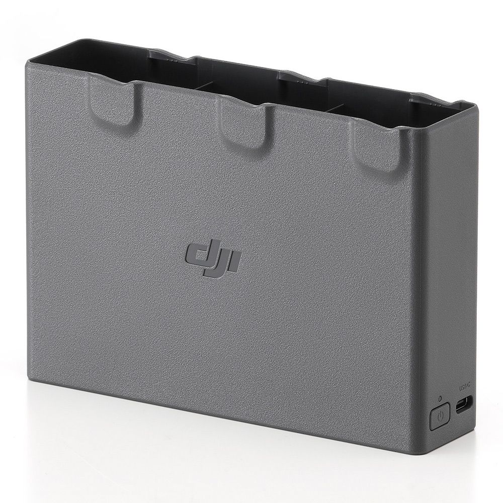 DJI Avata 2 Two-Way Charging Hub -lataustelakka on harmaa latauskeskitin, jossa on paikat kolmelle akulle, DJI:n logo etupuolella sekä USB-C-portti ja virtapainike sivussa, kuvassa valkoisella pinnalla.