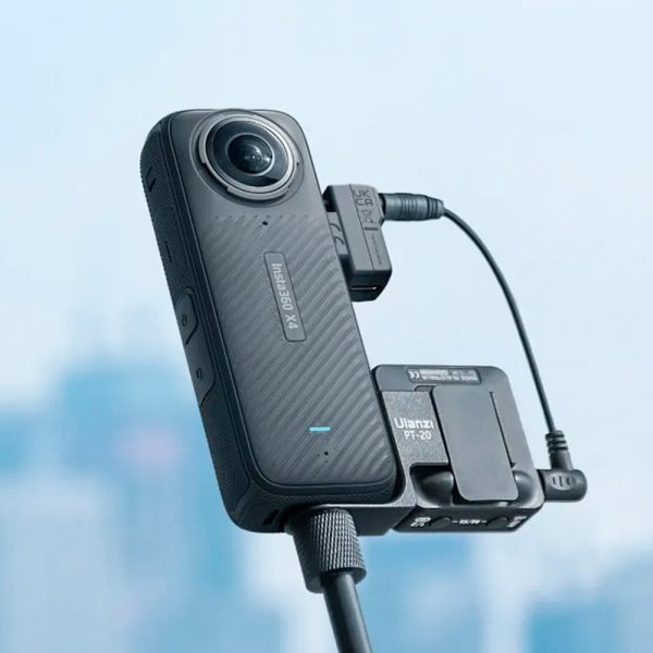 Lähikuva Insta360 X4 Mic Adapter -mikrofonisovittimesta, joka on liitetty mustaan 360-asteen toimintakameraan, joka on kiinnitetty jalustalle ja jossa on kaapelit. Sumealla taustalla näkyy sinisävyinen kaupunkikuva.