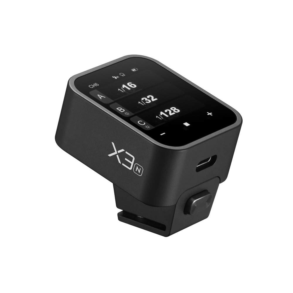 Pienikokoinen musta digitaalinen laite, jossa on merkintä "Godox X3 lähetin - Nikon" ja jossa on kosketusnäyttö, jossa on numeroita ja symboleja, USB-portti ja joka on suunniteltu kiinnitettäväksi muihin laitteisiin.