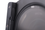 Lähikuva Nisi Filter Holder for Laowa 12mm f/2.8 -suodattimen pidikkeestä kiinnitettynä kameran objektiiviin, korostaen sen mustaa muotoilua, näkyviä ruuveja ja reunoja vaaleaa, neutraalia taustaa vasten.