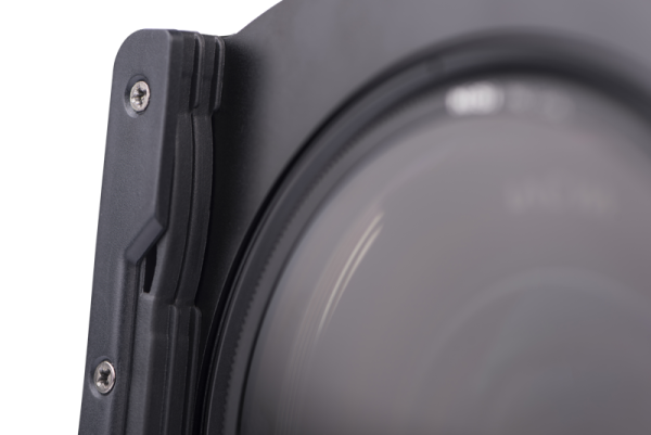 Lähikuva Nisi Filter Holder for Laowa 12mm f/2.8 -suodattimen pidikkeestä kiinnitettynä kameran objektiiviin, korostaen sen mustaa muotoilua, näkyviä ruuveja ja reunoja vaaleaa, neutraalia taustaa vasten.