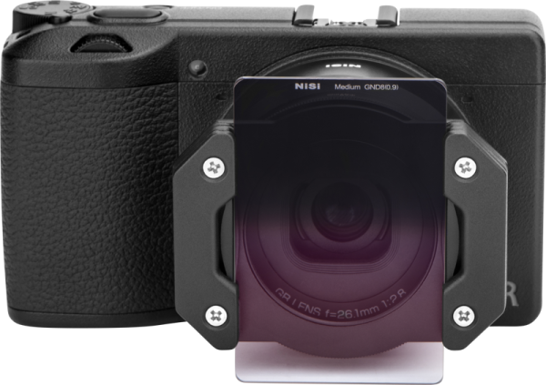Musta digitaalikamera, jossa on NiSi Master Kit For Ricoh GR IIIx, jonka etupuolelle on kiinnitetty suodatinpidike ja suorakulmainen gradienttilinssi-suodatin, kuvassa eteenpäin valkoista taustaa vasten.