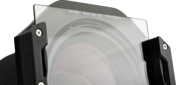 Close-up of a camera lens with a NiSi Square Black Mist 1/4 100x100mm filter mounted in front of it, held in a filter holder with metal screws on each side.