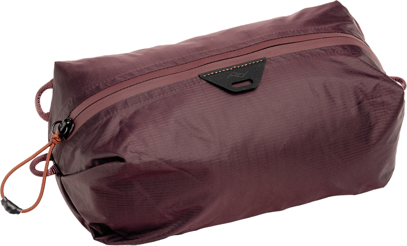 Peak Design Ultralight Packing Cube X-Small Eclipse on ruskeankeltainen, suorakulmainen pussi, jossa on musta kolmion muotoinen logo vetoketjun lähellä, oranssi naruveto, sivukahvat, ja se on valmistettu kevyestä, vedenkestävästä kankaasta.