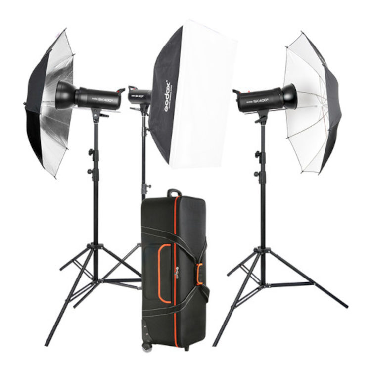 Godox SK400II Studio Flash Kit 400 D sisältää kolme studiovaloa, joissa on sateenvarjot ja softbox säädettävillä jalustoilla. Musta kantolaukku, jossa on oranssit korostukset, on sijoitettu valaisinasetelman eteen.