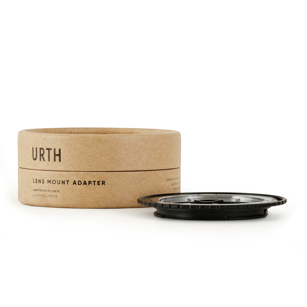 Pyöreä laatikko, jossa lukee "Urth Contax/Yashica (C/Y) - Canon (EF / EF S) objektiiviadapteri", on mustan metallisen objektiiviasennussovittimen vieressä valkoisella pohjalla.