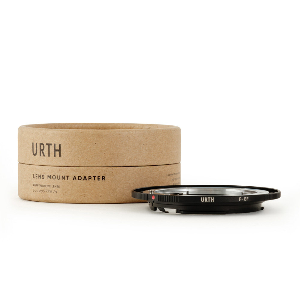 The black Urth Nikon F - Canon (EF / EF S) lens adapter is displayed next to its cylindrical brown cardboard packaging, which is marked URTH Lens Mount Adapter on a white background.