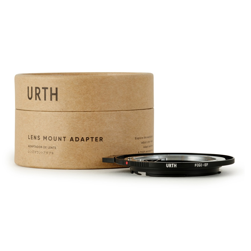 A cardboard box marked Urth Nikon F (G-type) - Canon (EF / EF S) lens adapter stands behind a black metal lens adapter ring marked URTH FJ(G)-EF against a white background.