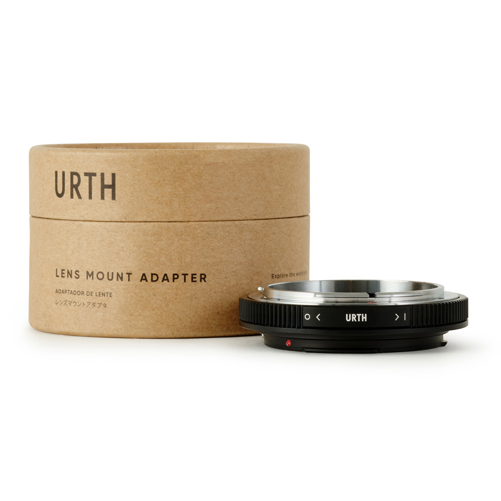 Lieriömäinen pahvilaatikko, jossa lukee Urth Canon FD - Canon (EF / EF S) objektiiviadapteri, on metallisen objektiiviasennussovittimen vieressä, jossa on musta harjattu reuna ja Urth-brändi painettuna siihen.