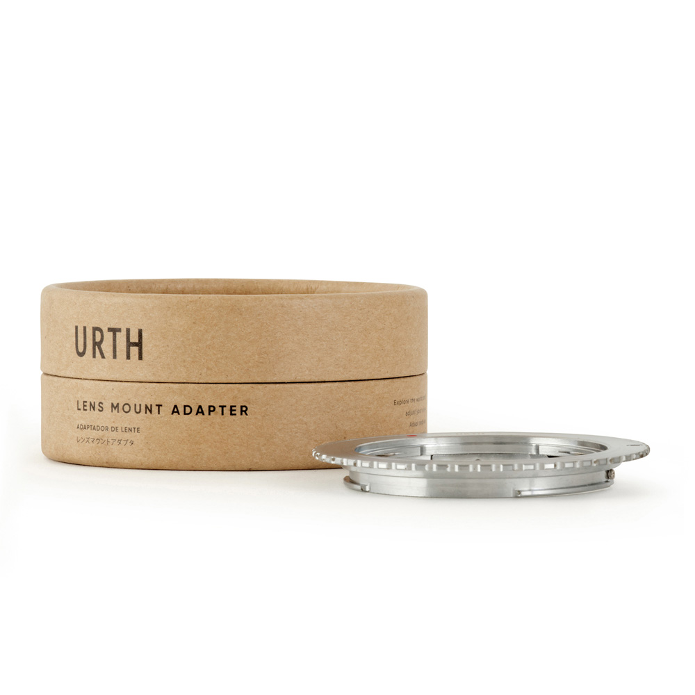 The cylindrical cardboard box, marked Urth Pentax K - Canon EF S lens adapter, is next to a silver, round metal lens mount adapter on a white background.