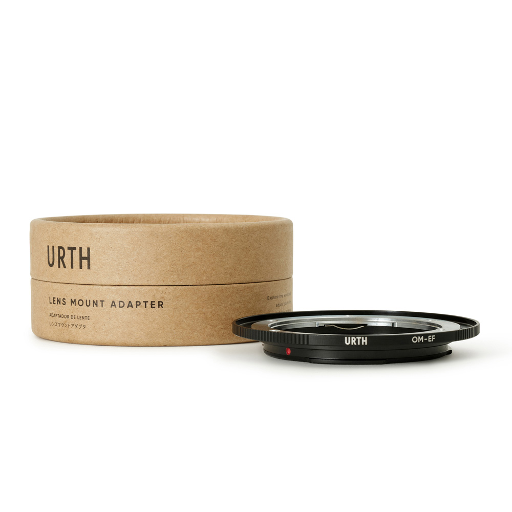 The Urth Olympus OM - Canon (EF/EF S) lens adapter sits next to its brown cylindrical box marked "URTH Lens Mount Adapter" against a white background.
