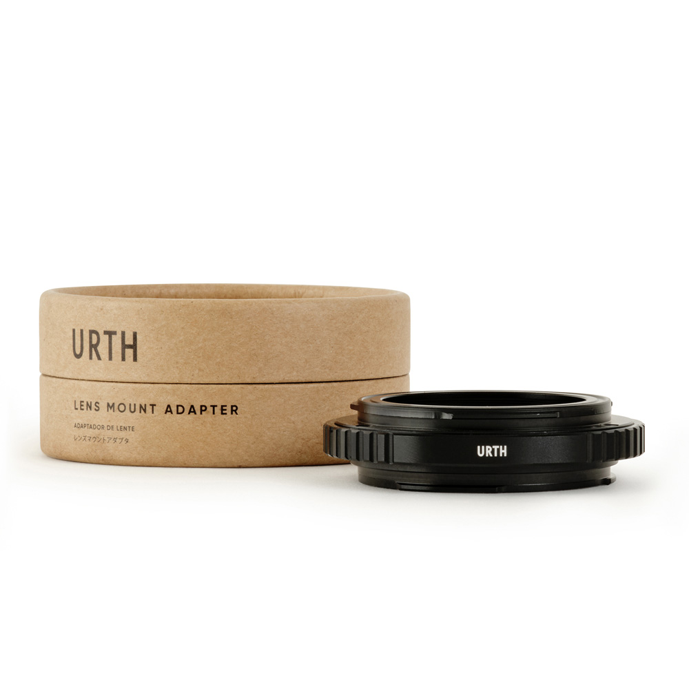 The black Urth Tamron T Mount to Canon (EF/EF S) lens adapter is placed in front of a round, brown cardboard box marked URTH Lens Mount Adapter on a white background.