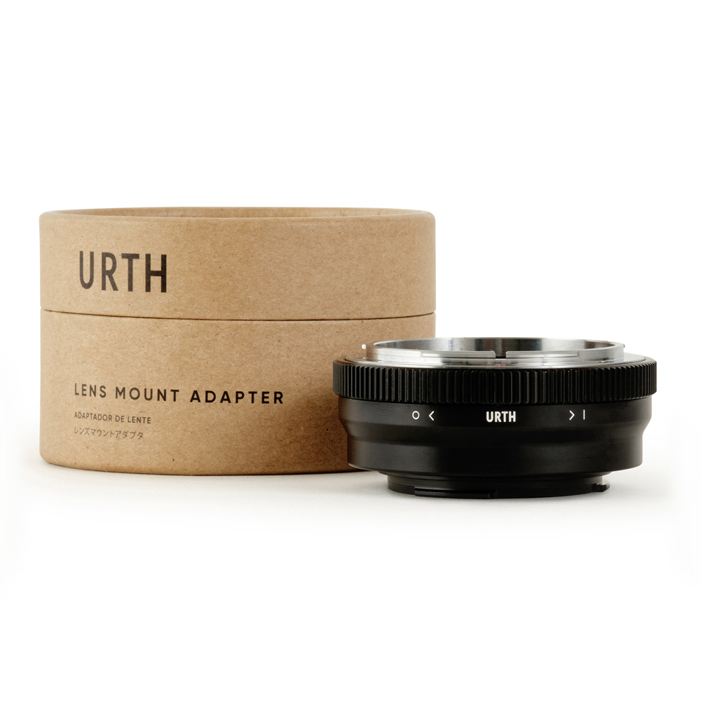 Musta Urth Canon FD - Canon EF M objektiiviadapteri on esillä sen lieriömäisen ruskean pahvilaatikon edessä, jossa lukee "URTH Lens Mount Adapter", ja molemmissa on metallisia ja harjattuja yksityiskohtia valkoisella pohjalla.