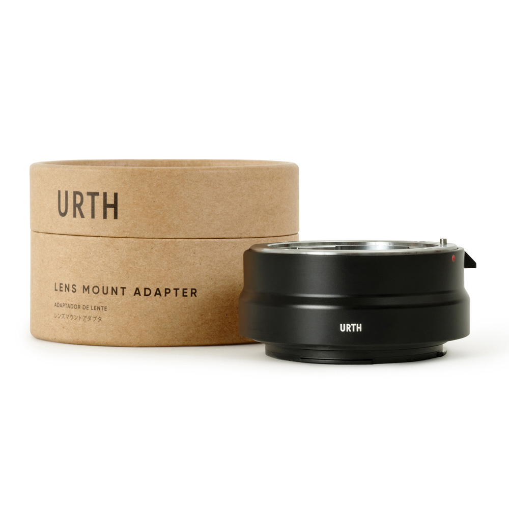 Musta Urth Pentax K - Canon RF objektiiviadapteri istuu sen lieriömäisen pahvilaatikon vieressä, jossa lukee URTH Lens Mount Adapteri valkoisella taustalla.