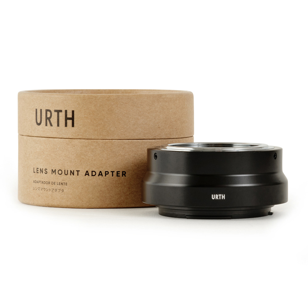 Musta Urth M42 - Canon RF objektiiviadapteri lepää sen lieriömäisen pahvipakkauksen vieressä, jossa lukee "URTH" ja "Lens Mount Adapter" mustalla tekstillä valkoisella pohjalla.