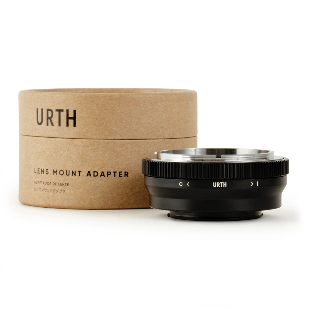 Musta Urth Canon FD - Fujifilm X objektiiviadapteri näkyy lieriömäisen ruskean laatikon edessä, jossa on merkintä URTH Lens Mount Adapter, jossa on näkyviä uria ja suuntausmerkintöjä.