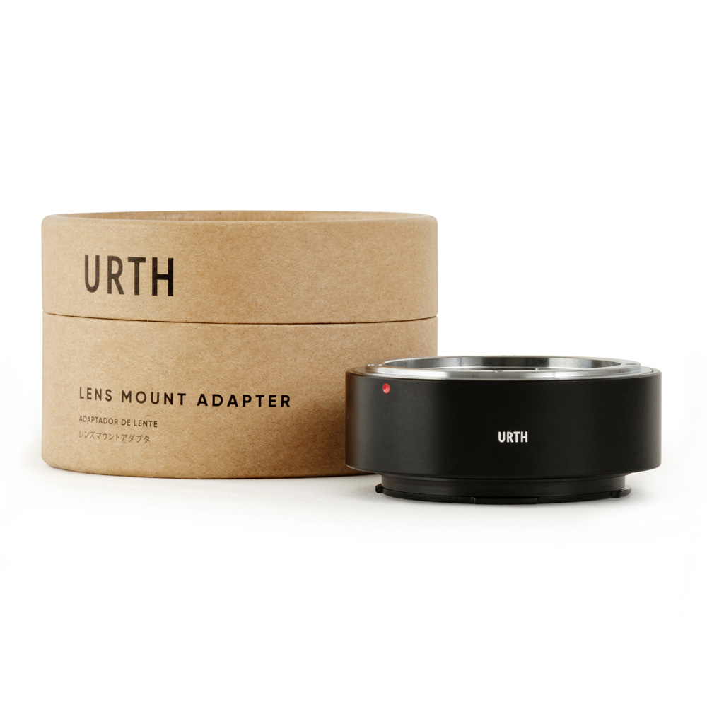 Musta Urth Konica AR - Leica L objektiiviadapteri näkyy sen lieriömäisen ruskean URTH-laatikon vieressä. Adapterissa on metallinen reunus ja pieni punainen piste.