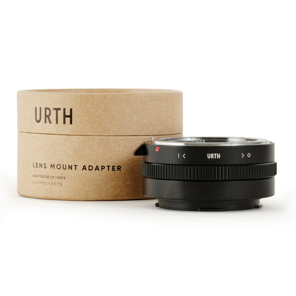Musta Urth Nikon F (G-tyyppi) - Leica L objektiiviadapteri istuu vieressä sen lieriömäinen ruskea pahvipakkaus merkitty URTH LENS MOUNT ADAPTER lihavoitu kirjaimilla.