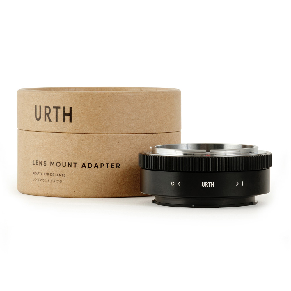 Lieriömäinen pahvilaatikko, jossa lukee "Urth Canon FD - Leica L objektiiviadapteri", on mustan metallisen objektiivisovittimen vieressä, jossa on valkoiset merkinnät ja Urth-logo, molemmat valkoista taustaa vasten.