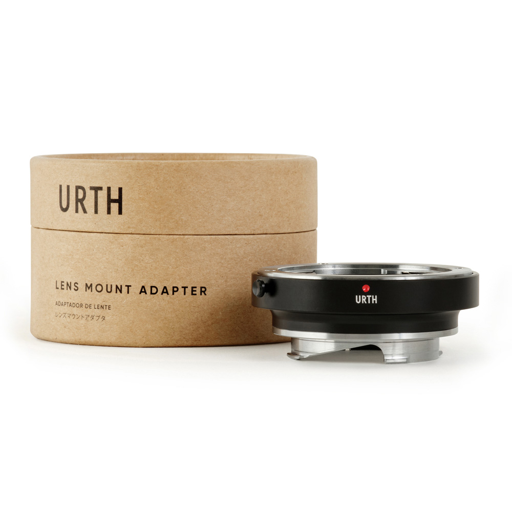 Urth Contax/Yashica (C/Y) - Leica M objektiiviadapteri näkyy lieriömäisen ruskean pakkauksen edessä, jossa lukee "URTH LENS MOUNT ADAPTER". Sovittimessa on musta ja hopeinen viimeistely, jossa on punainen kohdistuspiste.