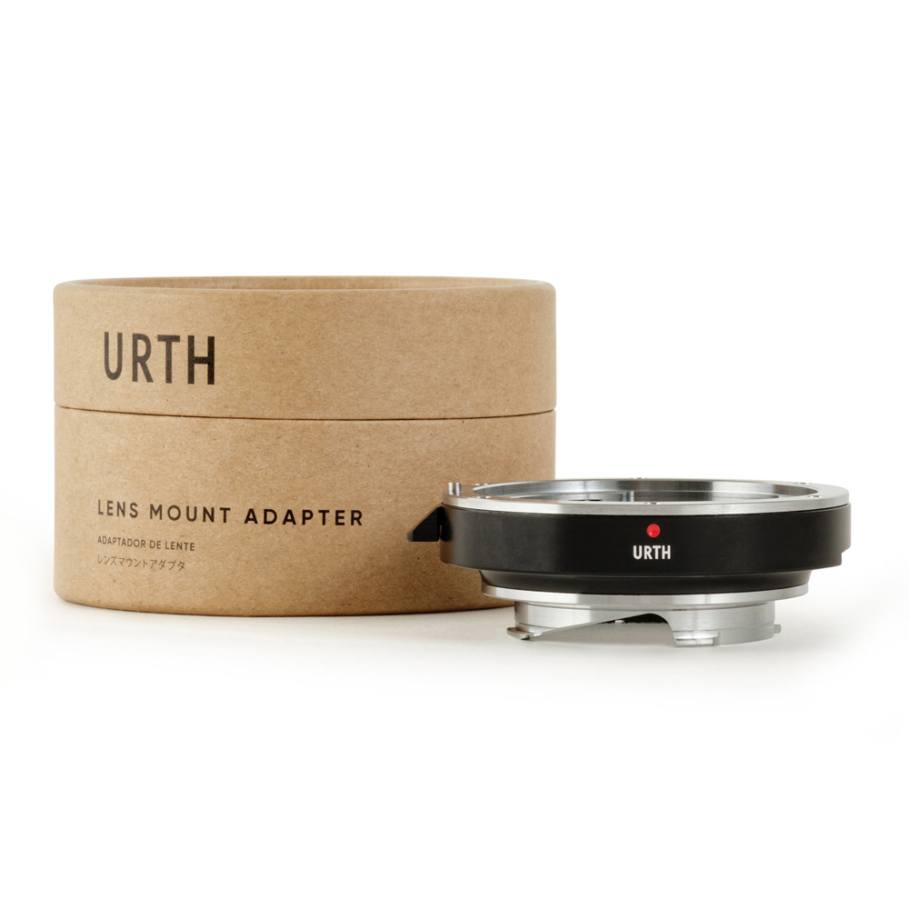Musta ja hopea Urth Canon (EF / EF S) - Leica M objektiiviadapteri näkyy sen lieriömäisen ruskean pahvilaatikon vieressä, jossa on merkintä URTH Lens Mount Adapter.