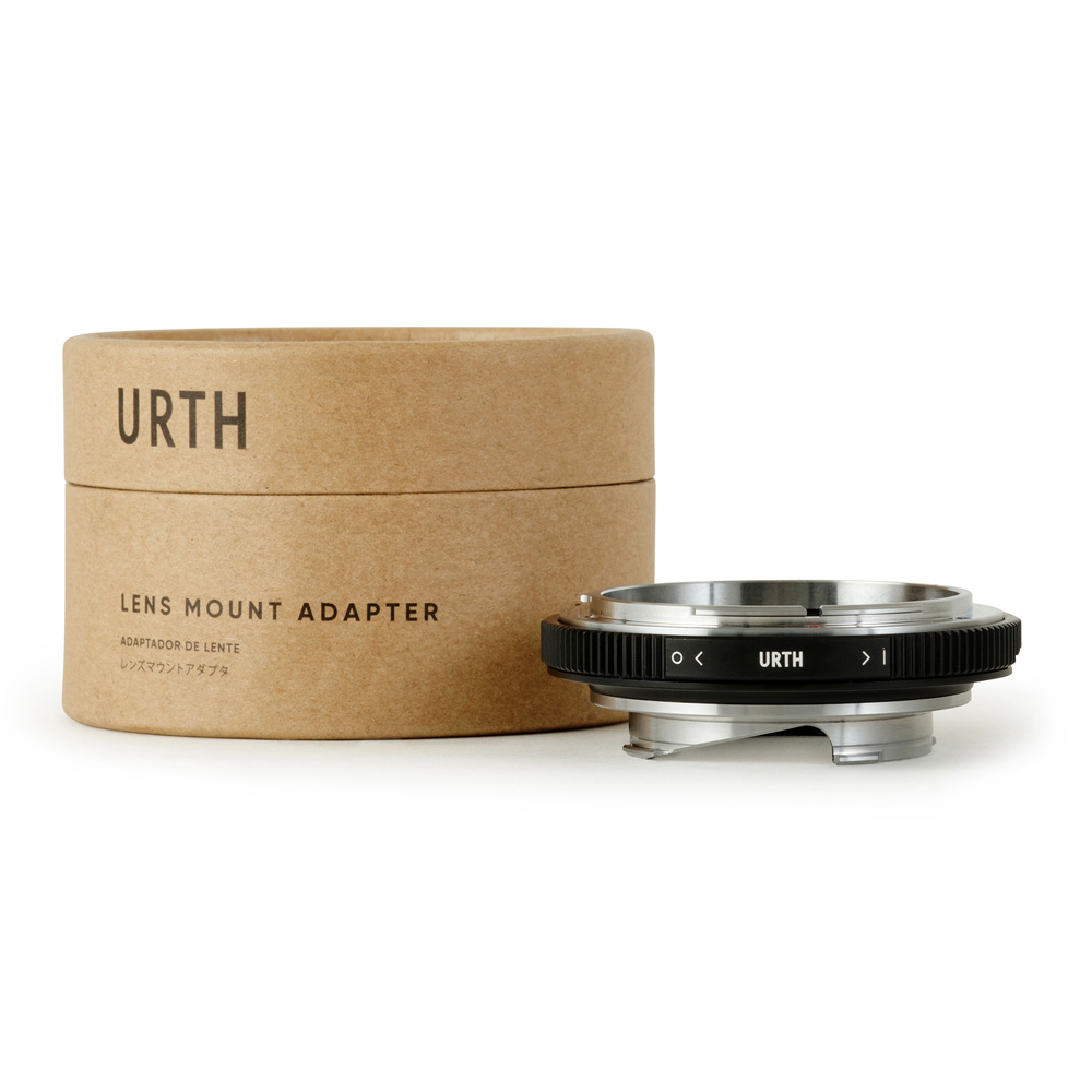 Sylinterimäinen pahvilaatikko, jossa lukee Urth Canon FD - Leica M objektiiviadapteri, on metallisen ja mustan objektiiviliitäntäsovittimen vieressä, johon on painettu URTH, ja molemmat on sijoitettu valkoiselle taustalle.