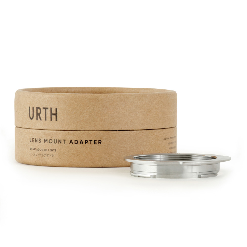 Pyöreä pahvilaatikko, jossa lukee "Urth M39 - Leica M objektiiviadapteri (28 90mm Frame Lines)", on metallisen objektiiviasennussovittimen vieressä valkoisella pohjalla.