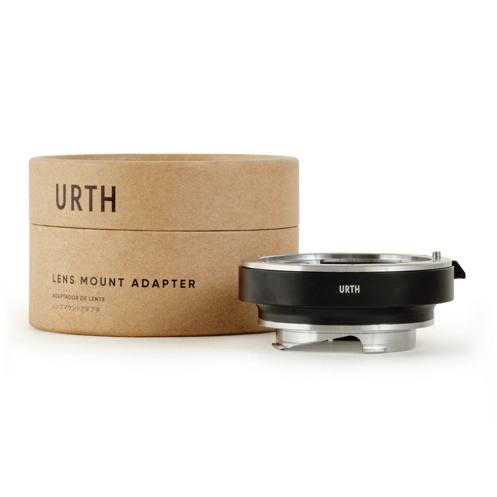 Lieriömäinen pahvilaatikko, jossa lukee "Urth Leica R - Leica M objektiiviadapteri", on metallisen ja mustan kameran objektiivin kiinnityssovittimen vieressä, molemmat valkoisella pohjalla.