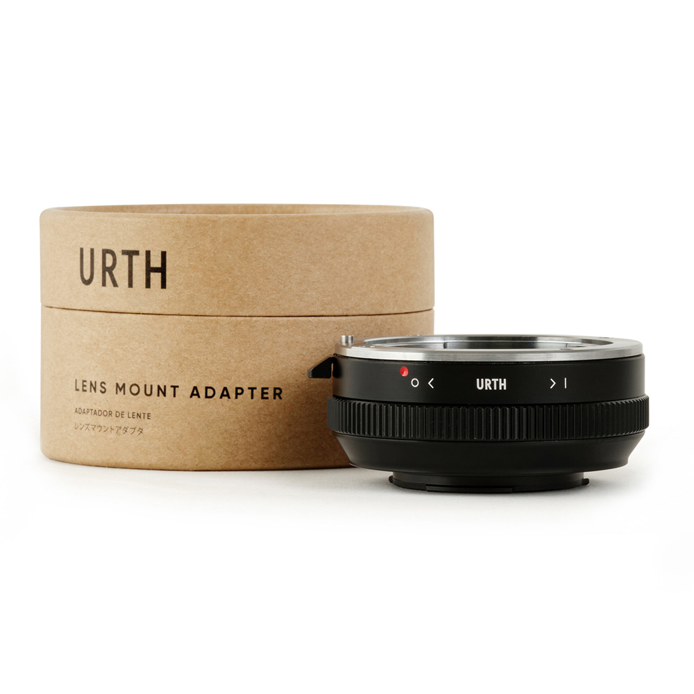 Musta Urth Sony A (Minolta AF) - Micro Four Thirds (M4/3) objektiiviadapteri istuu ruskean sylinterimäisen laatikon vieressä, jossa on merkintä URTH Lens Mount Adapter, valkoisella taustalla.