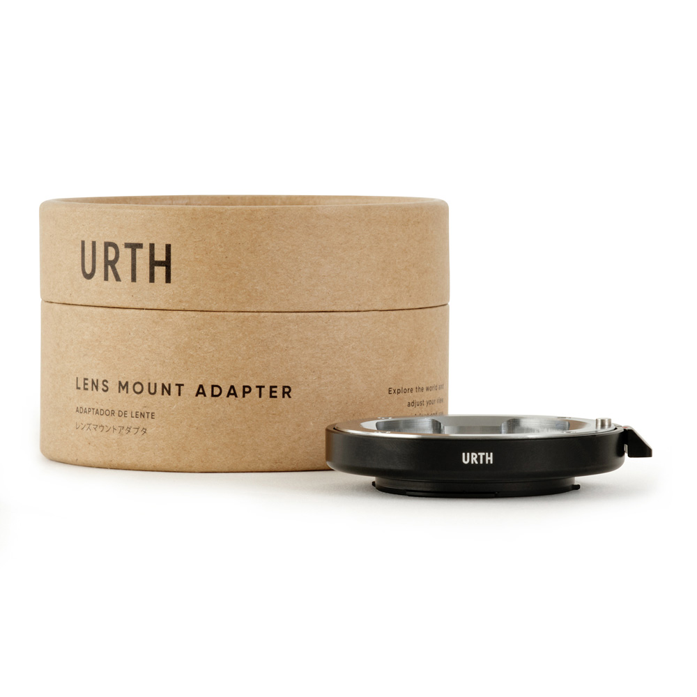 A cylindrical cardboard box with the words "Urth Leica M - Micro Four Thirds (M4/3) lens adapter" stands behind a black metal lens mount adapter, also marked URTH, on a white background.