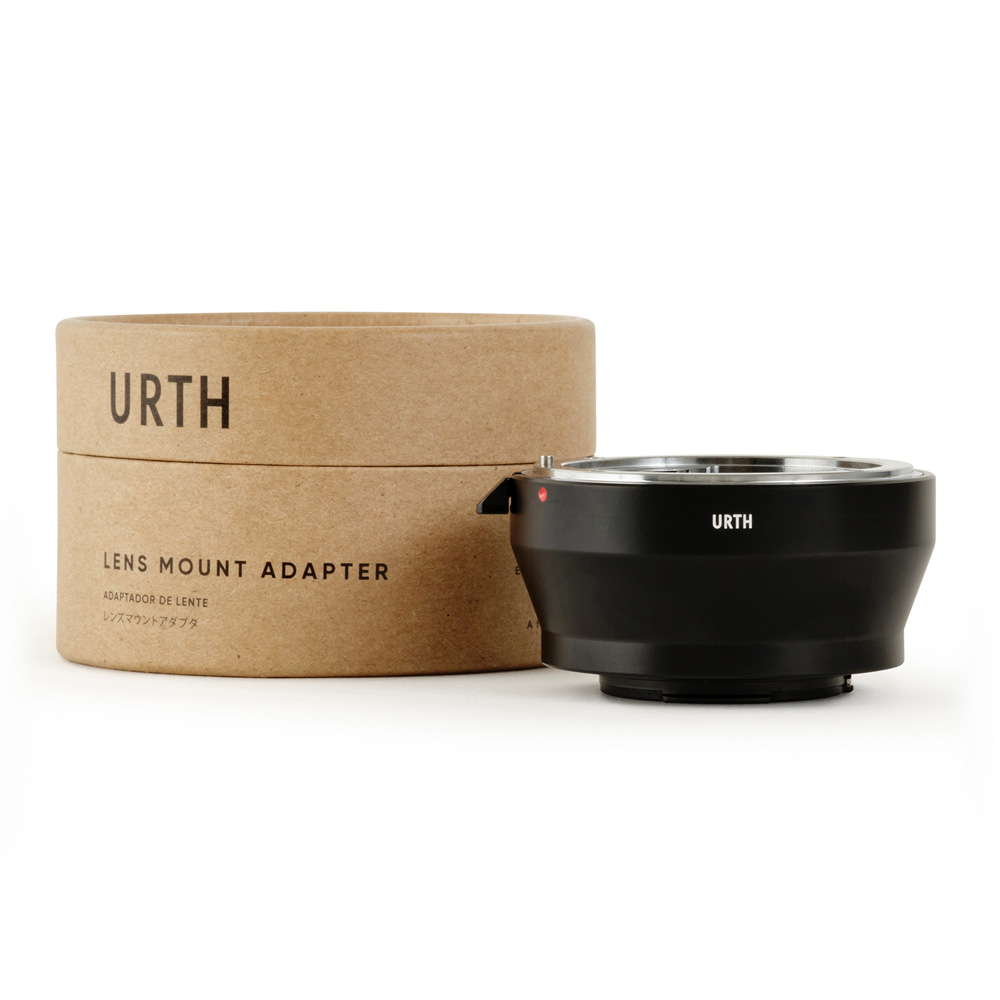 Musta Urth Nikon F - Nikon 1 objektiiviadapteri istuu sen lieriömäisen ruskean pahvipakkauksen vieressä, jossa lukee "URTH LENS MOUNT ADAPTER" valkoisella taustalla.