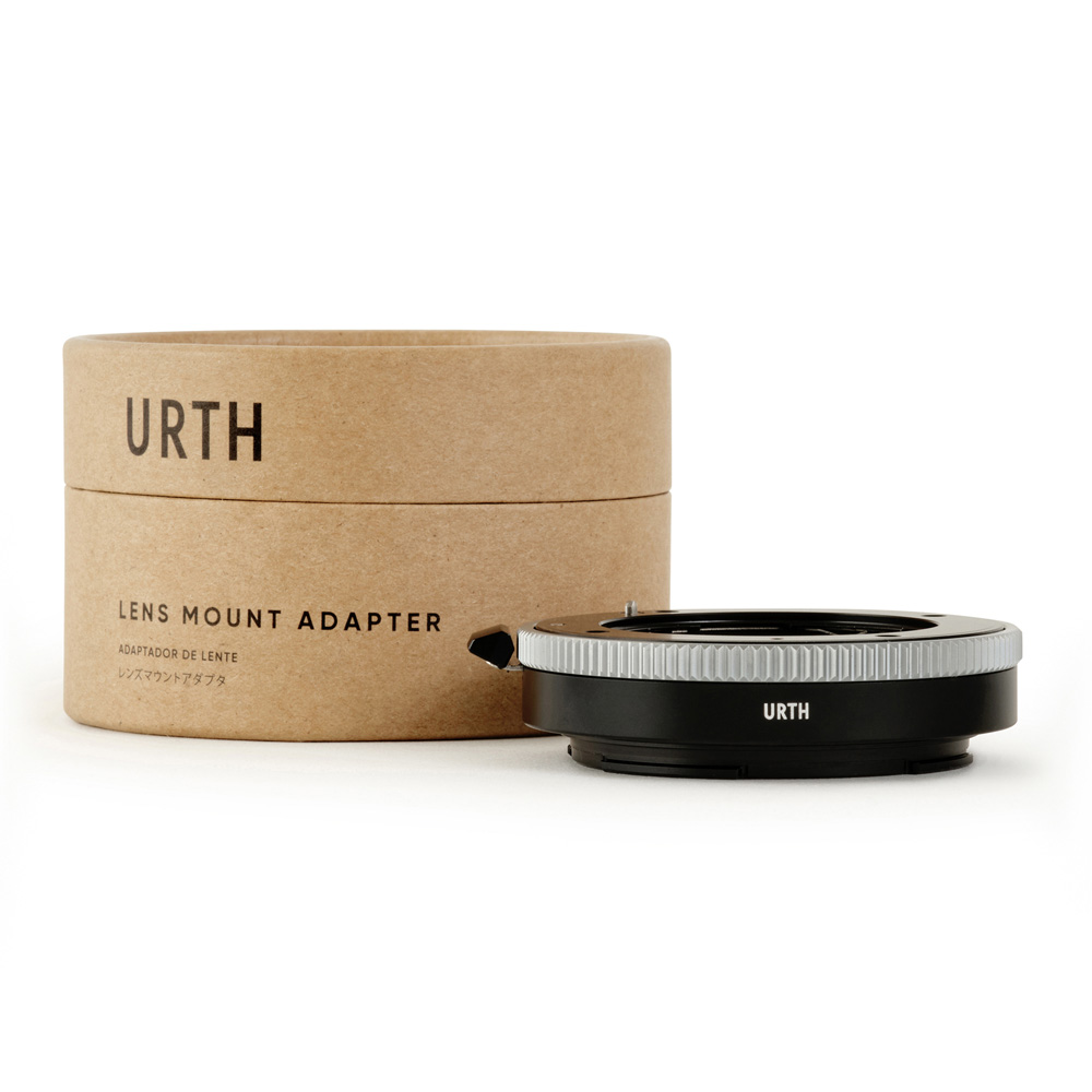 Urth Contax G - Nikon Z objektiiviadapteri näkyy sen ruskean lieriömäisen laatikon vieressä, jossa on merkintä URTH LENS MOUNT ADAPTER. Musta adapteri, jossa on hopeinen rengas, on sijoitettu pakkauksen eteen.