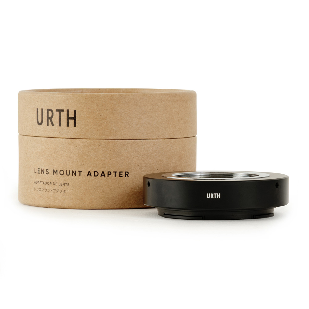 Pyöreä pahvilaatikko, jossa lukee "Urth M39 - Nikon Z objektiiviadapteri", on mustan metallisen objektiiviasennussovittimen vieressä, molemmat valkoisella pohjalla.