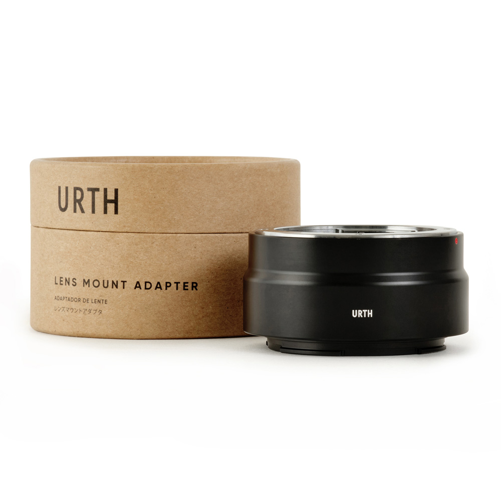 Musta Urth Olympus OM - Nikon Z objektiiviadapteri istuu sen ruskean sylinterimäisen pahvipakkauksen vieressä, jossa on merkintä URTH LENS MOUNT ADAPTERI valkoisella taustalla.