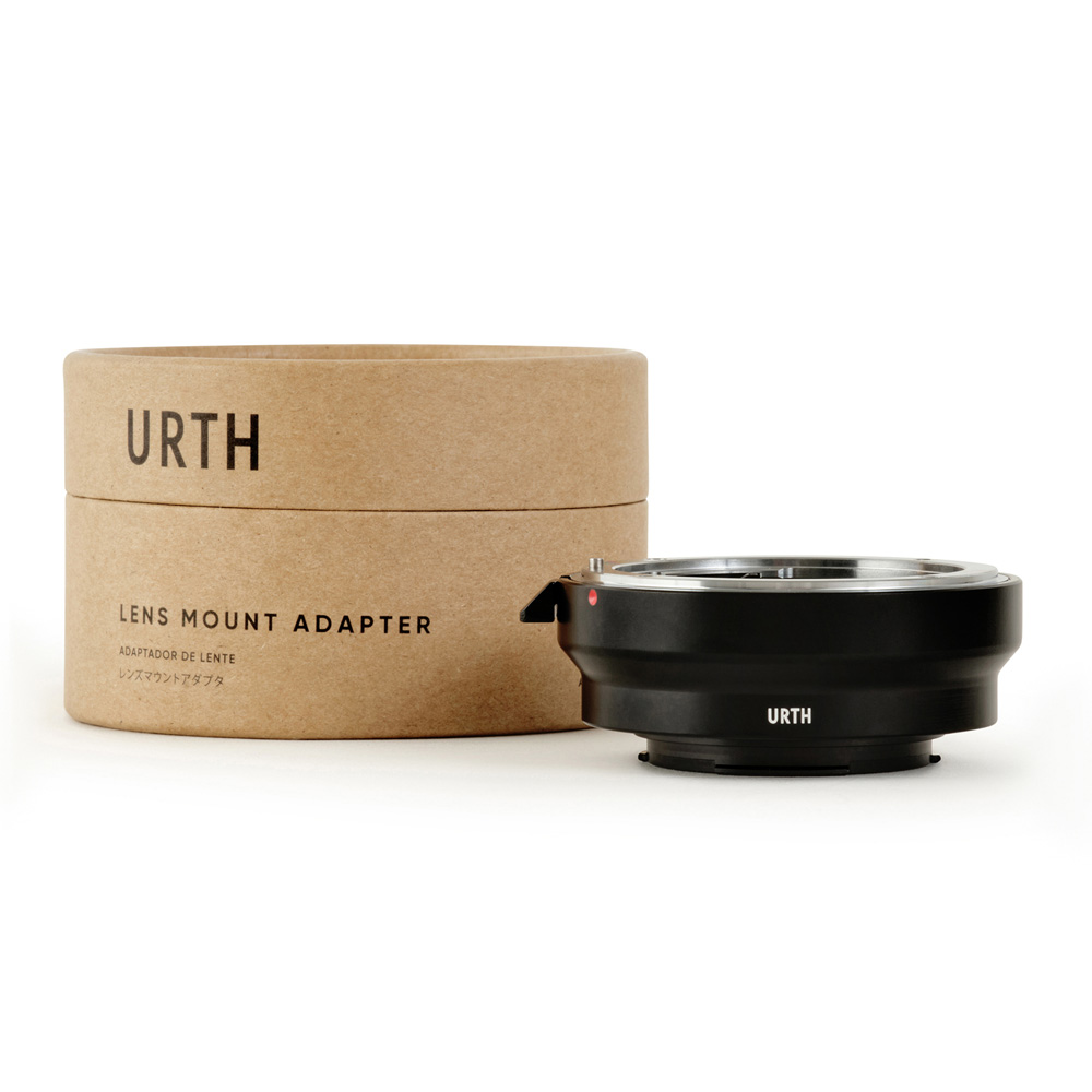 Urth Nikon F - Samsung NX objektiiviadapteri on sijoitettu sen sylinterimäisen ruskean pahvipakkauksen viereen, jossa lukee mustalla "URTH Lens Mount Adapter". Adapteri on musta ja siinä on hopeinen sisärengas.