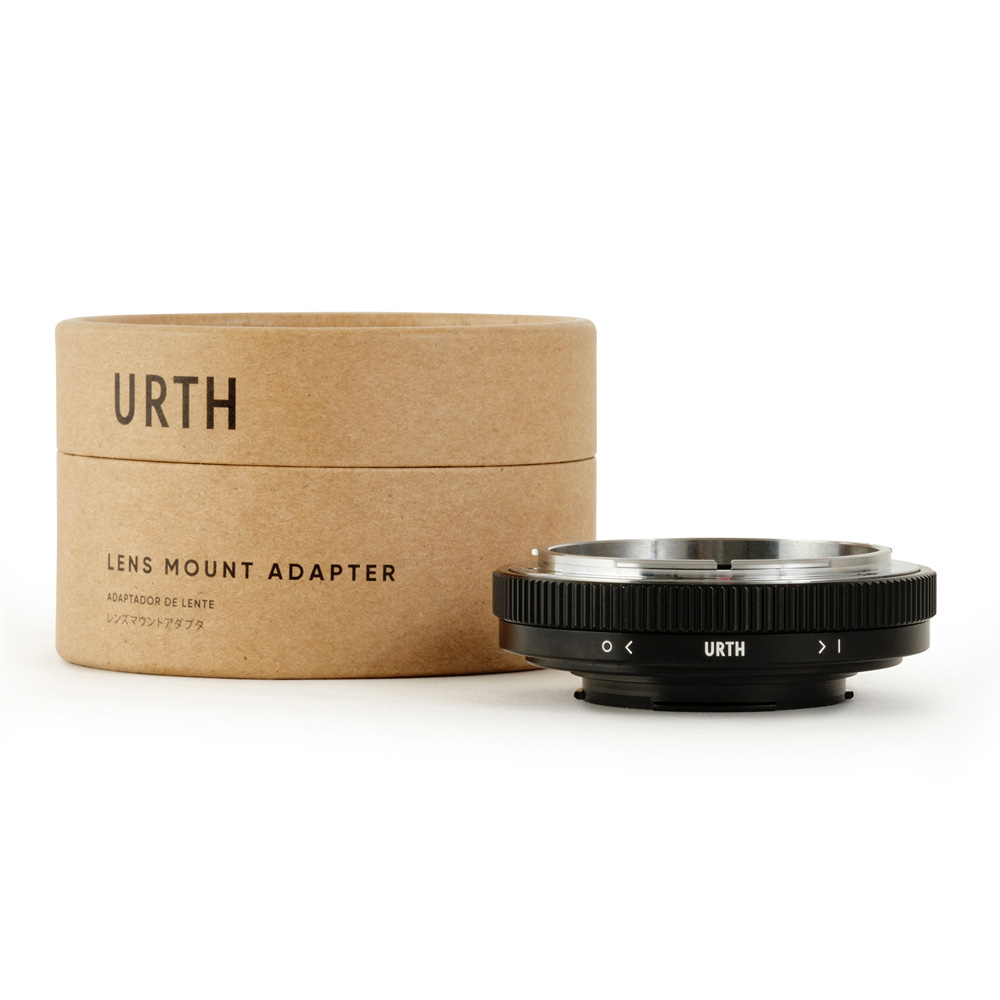 Pyöreä pahvilaatikko, jossa lukee "Urth Canon FD - Samsung NX objektiiviadapteri", seisoo mustan metallisen objektiivikiinnityssovittimen vieressä, jossa on URTH-logo, ja molemmat näkyvät valkoisella pohjalla.